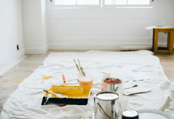 Paint trays, brushes and rollers on a protective sheet spread over the floor of a bright room