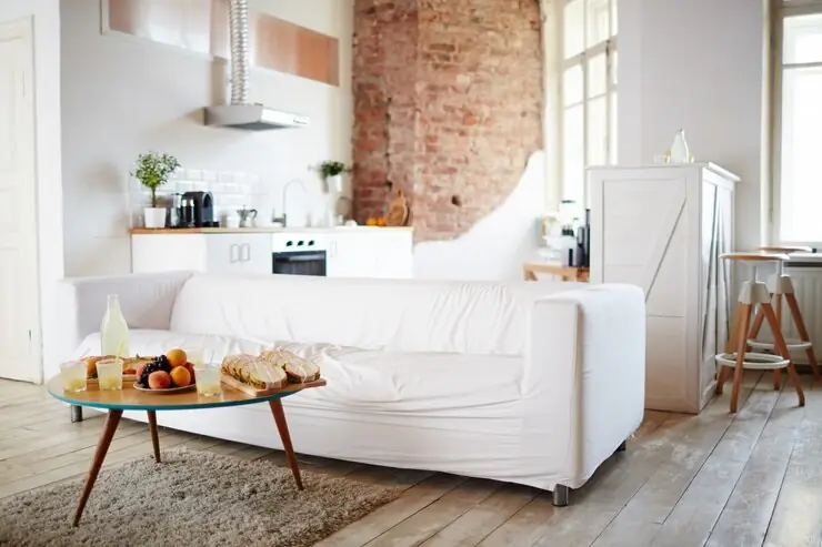 A furnished living room with a white sofa, a brick wall and an open kitchen